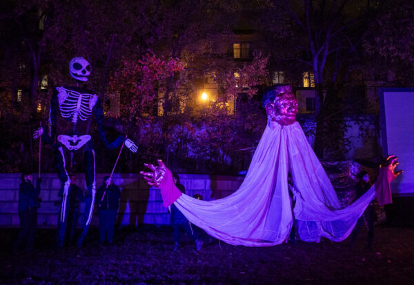 A roughly 16 foot puppet, with gold head and hands, and white fabric draping for body is puppetted on the grass next to a roughly 20 foot skeleton puppet with wings.