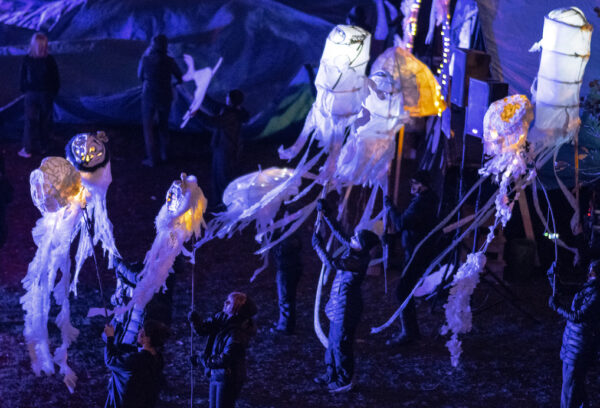 Nine people in black walking at night with lit up white paper and fabric lanterns and puppet heads on long sticks.