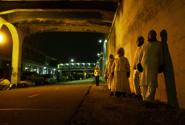 Four people dressed in white robes, standing under a city bridge. One other person not in all white is standing behind them.