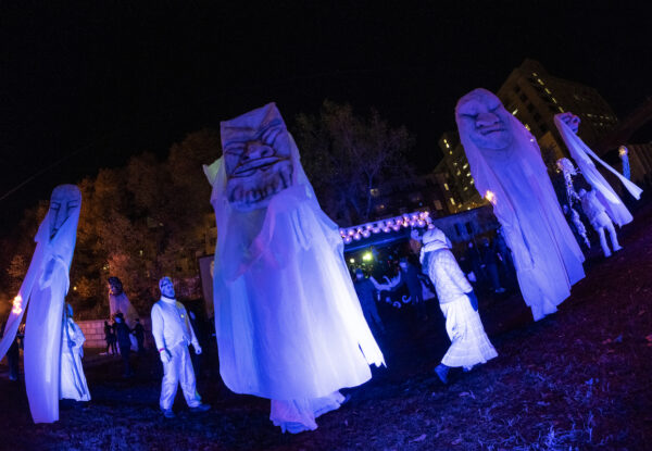 a couple people dressed in white robes, and to people holding large white puppet heads atop long poles with white fabric robes descending beneath the heads and covering the puppeteers.