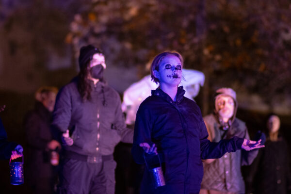 Several people dressed in black with skull face makeup, holding unlit canister lanterns.