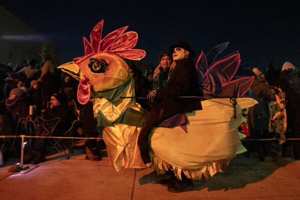 A man dressed in black with skull face makeup sitting in the middle of a 5 foot tall, and 5 - 6 foot long rooster puppet.