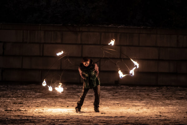 A person dressed in black, outdoors in the snow, with metal bat wings on their back that have the edges lit on fire.