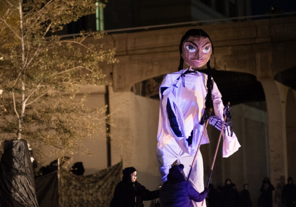 A couple puppeteers, outdoors, dressed in black, puppetting (from the ground) a 20 foot tall puppet of a girl with black hair in a white outfit.
