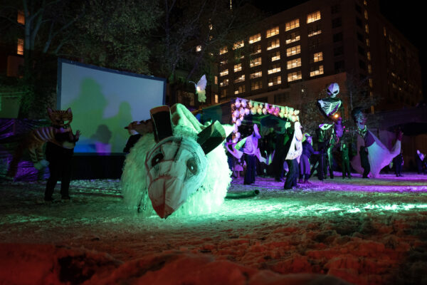 A four foot tall, opposum puppet, on the snowy ground, with performers, dressed in back, and a building in the background.