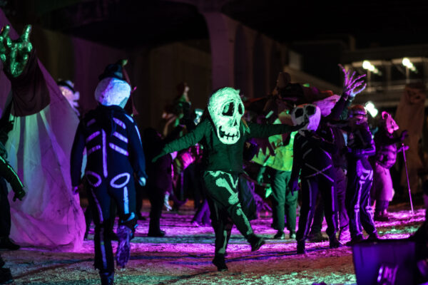 Several performers dressed in black, dancing and wearing large paper mache skull heads.