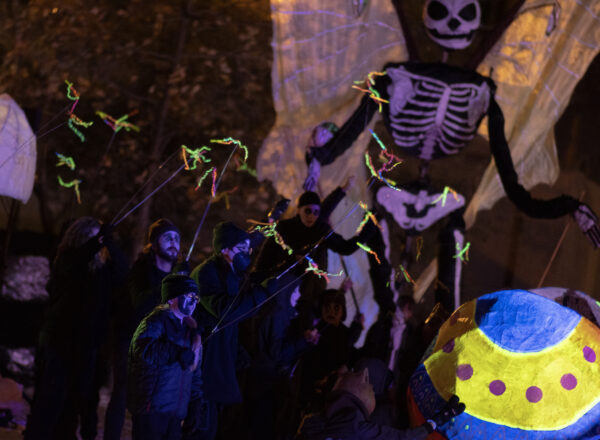 People, dressed in black, waving long thin poles with neon colored thin scraps at the end, with a 20 foot skeleton puppet in the background.