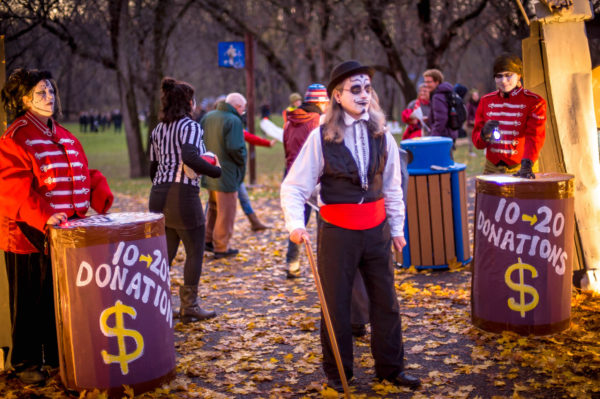 A wooded park in autumn (golden leaves on the ground). There are two almost three foot high round tables on either side of an entry gate. The bottoms are covered in paper with the words $10-$20 Donations. At each table there is a person with skull face makeup and a read band jacket. There is a greeter in front of them, wearing skull face makeup, black trousers, a black vest, ruffled white shirt and red cummerbund. He is also holding a cane. There are people walking around in the background.  