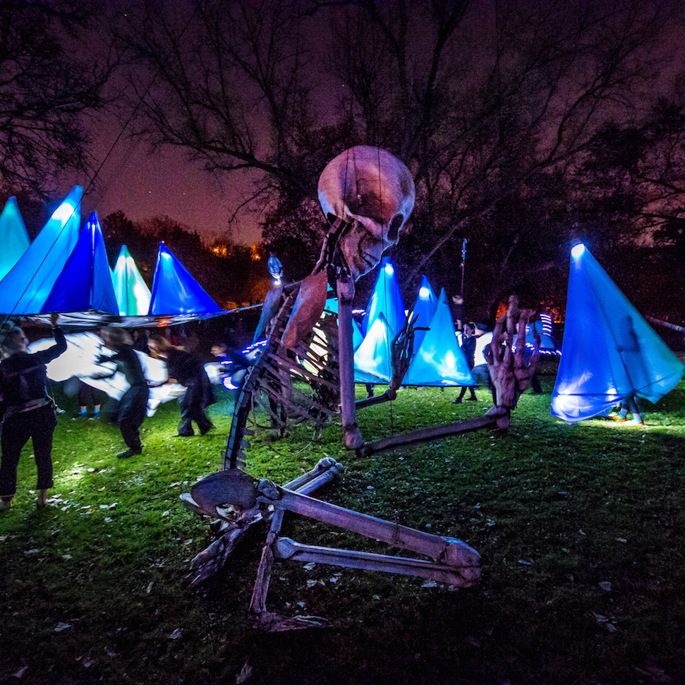 A 16 foot puppet skeleton kneeling on the ground with lighted triangle tents in the background.