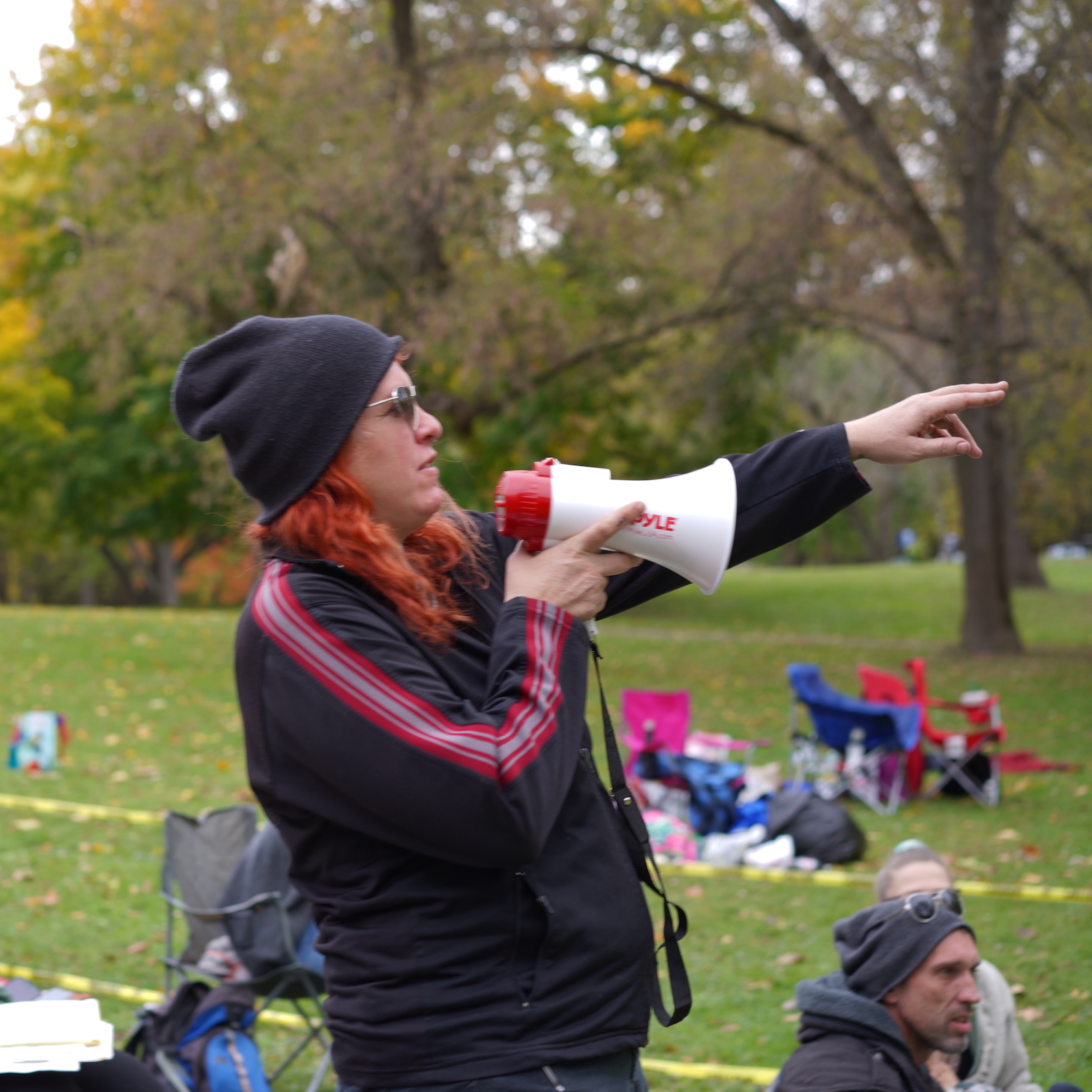A woman in a park speaking into a megaphone and pointing, with people in the background.